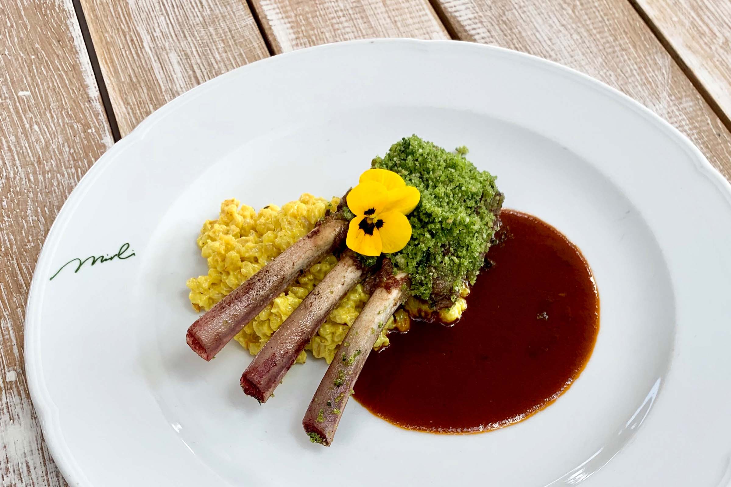 Ein Gourmetgericht mit drei Lammkoteletts mit einer Kruste aus grünen Kräutern, serviert mit cremigem gelbem Risotto, einer Lache reichhaltiger brauner Soße und garniert mit einer essbaren gelben Blume auf einem weißen Teller.